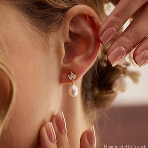 May include: A close-up of a gold earring with a teardrop pearl and small clear crystals. The earring is worn on an ear. The earring has a small floral design at the top. The background is blurred.