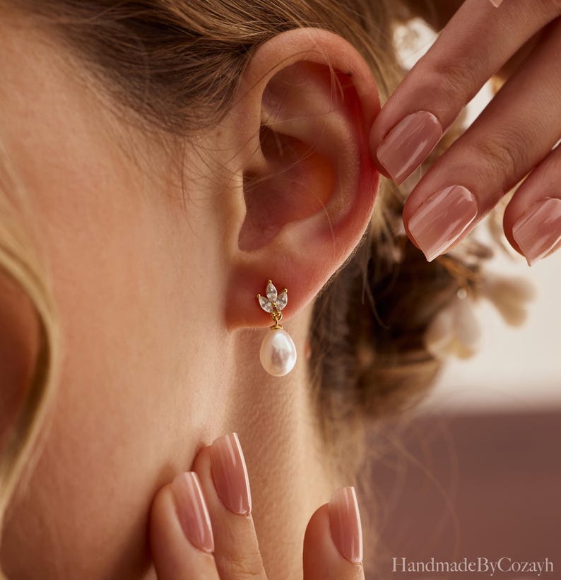 May include: A close-up of a gold earring with a teardrop pearl and small clear crystals. The earring is worn on an ear. The earring has a small floral design at the top. The background is blurred.