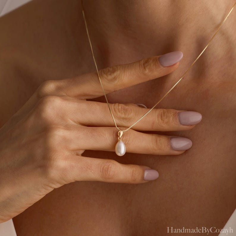May include: A delicate gold chain necklace featuring a single, teardrop-shaped white pearl pendant. The pendant is suspended from a simple gold bail, creating a refined and understated piece of jewellery that rests against the collarbone area.