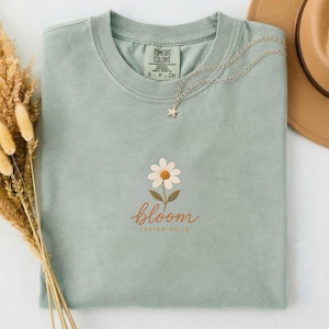 Puede incluir: Una camiseta verde salvia con una margarita blanca bordada con centro amarillo y hojas verdes. Debajo de la flor, la palabra bloom está bordada en cursiva naranja, seguida del texto ISAIAH 43:19 en letras naranjas más pequeñas.