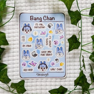 May include: A rectangular sticker sheet titled Bang Chan featuring multiple small illustrations of a blue-haired wolf character, trains, laptops, and musical notes. Text includes "IT'S FOR MOTIVATION," "IT'S ABOUT TRAINS," "WHY YOU MAD BRUH?," "BIG HUG!," and "WOLF CHAN."