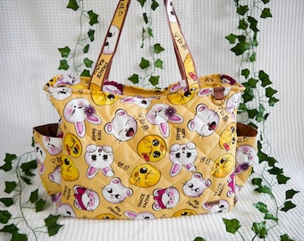 May include: Yellow quilted tote bag with a repeating pattern of cartoon characters, including bunnies and bears. The bag has brown accents, side pockets, and straps with text. The bag is decorated with the text "Stray Kids" and "Dance Racha".