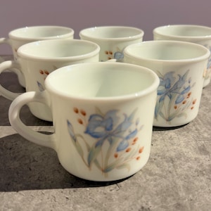 Könnte beinhalten: Sechs cremefarbene Keramik-Kaffeetassen mit Henkeln. Jede Tasse zeigt ein Blumendesign mit blauen Schwertlilien und kleinen orange-braunen Beeren. Die Tassen stehen auf einer grauen Oberfläche.