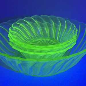 May include: A collection of vintage green glass bowls. The set comprises a large serving bowl and several smaller bowls nested within. The glass features a swirled pattern. The bowls glow brightly under a black light, highlighting their unique colour.