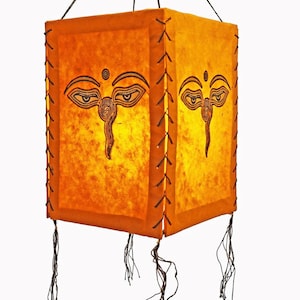 May include: A square, yellow paper lantern with a black Buddha eye design. The lantern has a fringe trim and is hanging from a string.