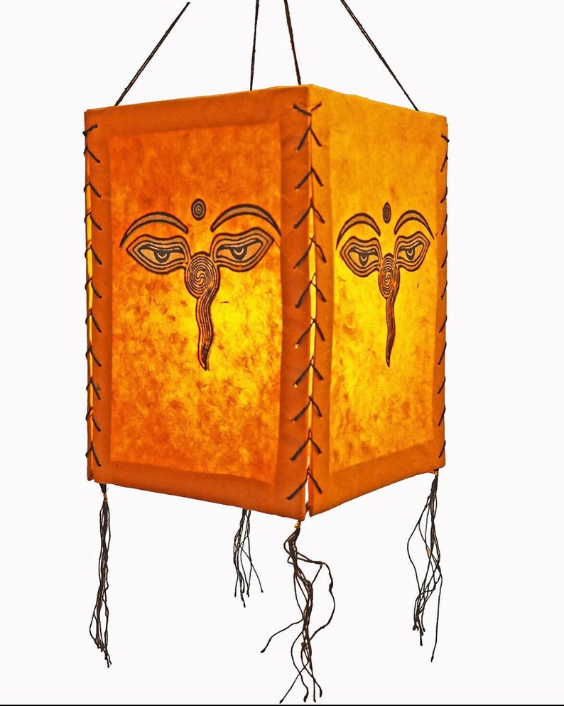 May include: A square, yellow paper lantern with a black Buddha eye design. The lantern has a fringe trim and is hanging from a string.