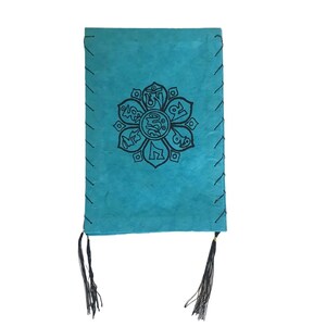 May include: A turquoise blue leather journal with a black mandala design and black tassels. The mandala features a flower with eight petals and Tibetan script.