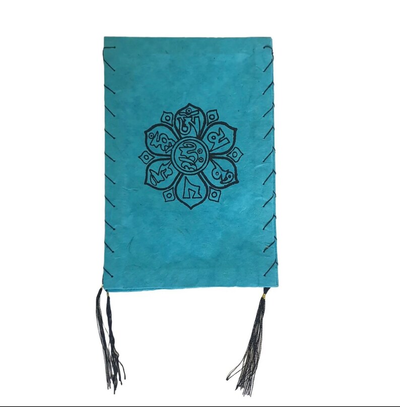 May include: A turquoise blue leather journal with a black mandala design and black tassels. The mandala features a flower with eight petals and Tibetan script.