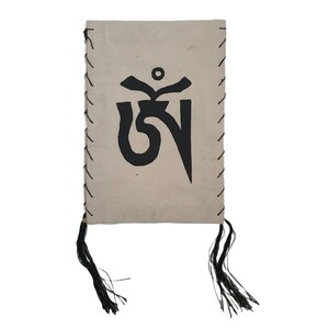 May include: A small, beige book with a black Tibetan prayer flag symbol printed on the front. The book has black tassels on the bottom.