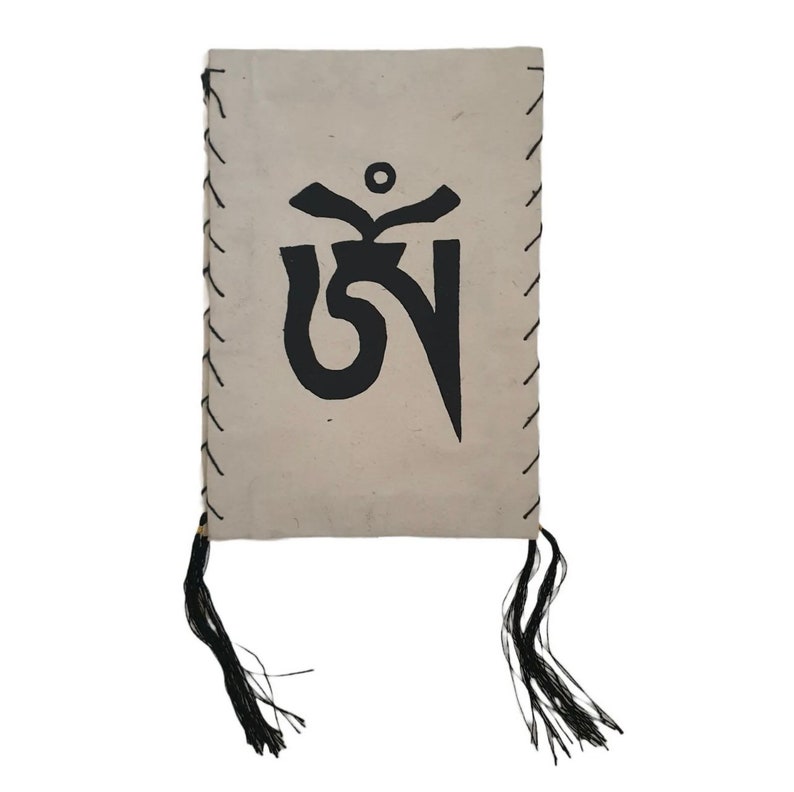 May include: A small, beige book with a black Tibetan prayer flag symbol printed on the front. The book has black tassels on the bottom.