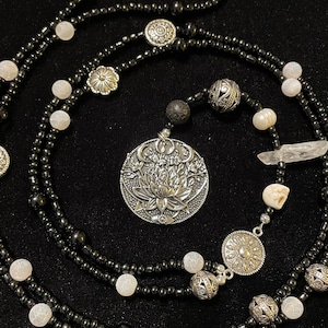 May include: A multi-strand necklace with black beads, white beads, and silver charms. The necklace features a large silver pendant with a floral design and a smaller silver pendant with a sun design. The necklace also includes a white crystal and a small white skull charm.