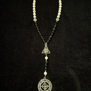 May include: A black and white beaded necklace with silver Celtic knot charms. The necklace has a triangular charm at the top and a round charm at the bottom.
