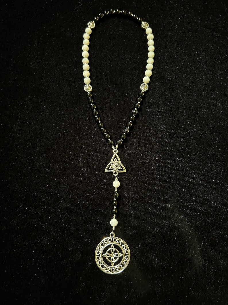 May include: A black and white beaded necklace with silver Celtic knot charms. The necklace has a triangular charm at the top and a round charm at the bottom.