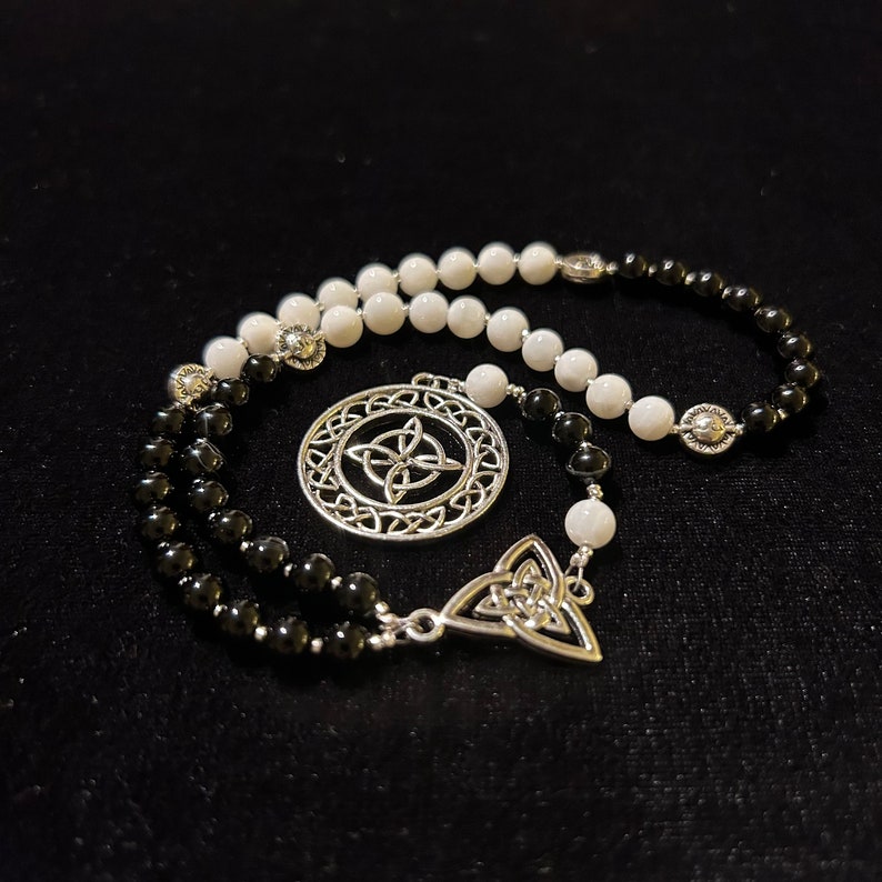 May include: A black and white beaded rosary with a silver Celtic knot pendant. The rosary has a circular pendant with a Celtic knot design and a smaller triangular pendant with a Celtic knot design.