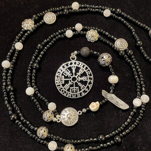 May include: A black and white beaded necklace with a silver pendant featuring a circular design with runes. The necklace also includes a silver skull bead, a clear quartz crystal, and other silver beads.