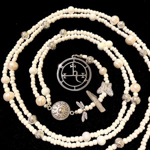 May include: White beaded necklace with a silver pendant featuring a symbol with a cross and a crescent moon. The necklace also includes a silver dragonfly charm and a silver butterfly charm.