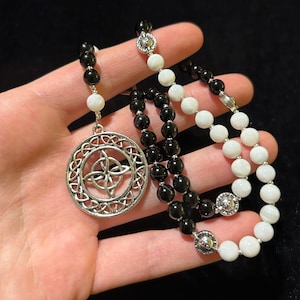 May include: A black and white beaded necklace with a silver Celtic knot pendant. The necklace is made with black and white beads and has a silver charm with a Celtic knot design.
