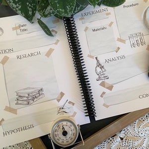 May include: A spiral-bound science journal with a magnifying glass and a stopwatch. The journal pages feature labeled sections for experiments, including "Question," "Research," and "Analysis." The journal is decorated with illustrations of scientific tools.