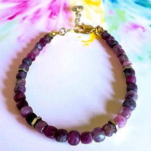 May include: A beaded bracelet featuring faceted, deep pink and purple gemstones. The bracelet is strung on a gold-colored chain and has gold-colored metal accents. The background is a soft, colorful, abstract design.