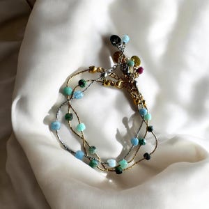 Könnte beinhalten: Ein mehrsträngiges Armband mit Gold- und Silberketten. Das Armband hat verschiedene Perlen in Blau-, Grün- und Goldtönen. Kleine Charms und Akzente sind ebenfalls sichtbar.