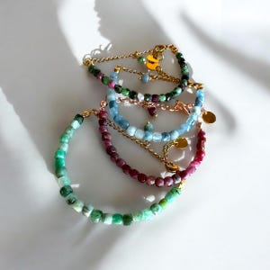 Könnte beinhalten: Vier Armbänder mit verschiedenen Edelsteinen in verschiedenen Farben und goldenen Akzenten. Die Armbänder sind übereinander gestapelt. Das oberste Armband hat grüne Perlen, das zweite Armband hat blaue Perlen, das dritte Armband hat rosa Perlen und das unterste Armband hat grüne Perlen.