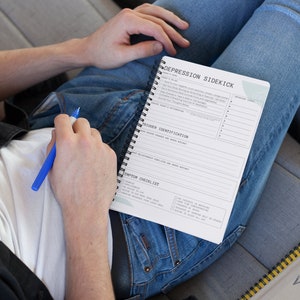 Può includere: Un quaderno a spirale bianco e grigio con il titolo "DEPRESSION SIDEKICK" e un elenco di controllo intitolato "SYMPTOM CHECKLIST". Il quaderno è aperto a una pagina con un elenco di sintomi che includono "SADNESS", "HOPELESSNESS", "LOSS OF INTEREST", "DIFFICULTY SLEEPING", "CHANGES IN APPETITE", "FEELING WORTHLESS", "DIFFICULTY CONCENTRATING", "RECURRING THOUGHTS OF DEATH", "DESIRE TO HARM SELF OR OTHERS", "DESIRE TO SLEEP ALL DAY".