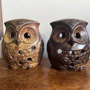 May include: Two ceramic owl figurines featuring a textured, speckled glaze. The left owl is light brown with darker accents, while the right owl is dark brown. Both figures have circular cutouts on their chests and faces, creating a hollow design.
