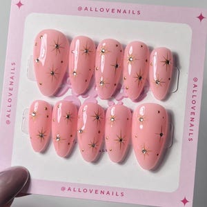 May include: A set of ten pink press-on nails with a starburst and dot design. Each nail features a gold starburst and small gold dots, with a few nails also having a small gem. The nails are displayed on a pink card with the text "@ALLOVENAILS".