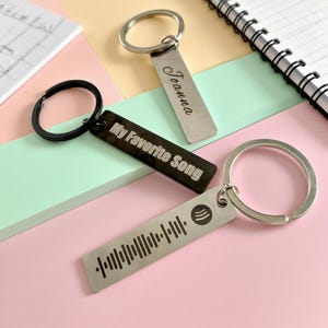 May include: Three keyrings with rectangular metal tags and silver key rings. One black tag reads "My Favorite Song" in white. Another silver tag is engraved with the name "Joanna". The third keyring has a Spotify code.