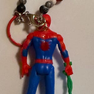 May include: A Spiderman figurine with a beaded lanyard that spells "SPIDEY". The figurine is red and blue, holding a green web-like accessory. The lanyard has blue, white, red, and black beads.