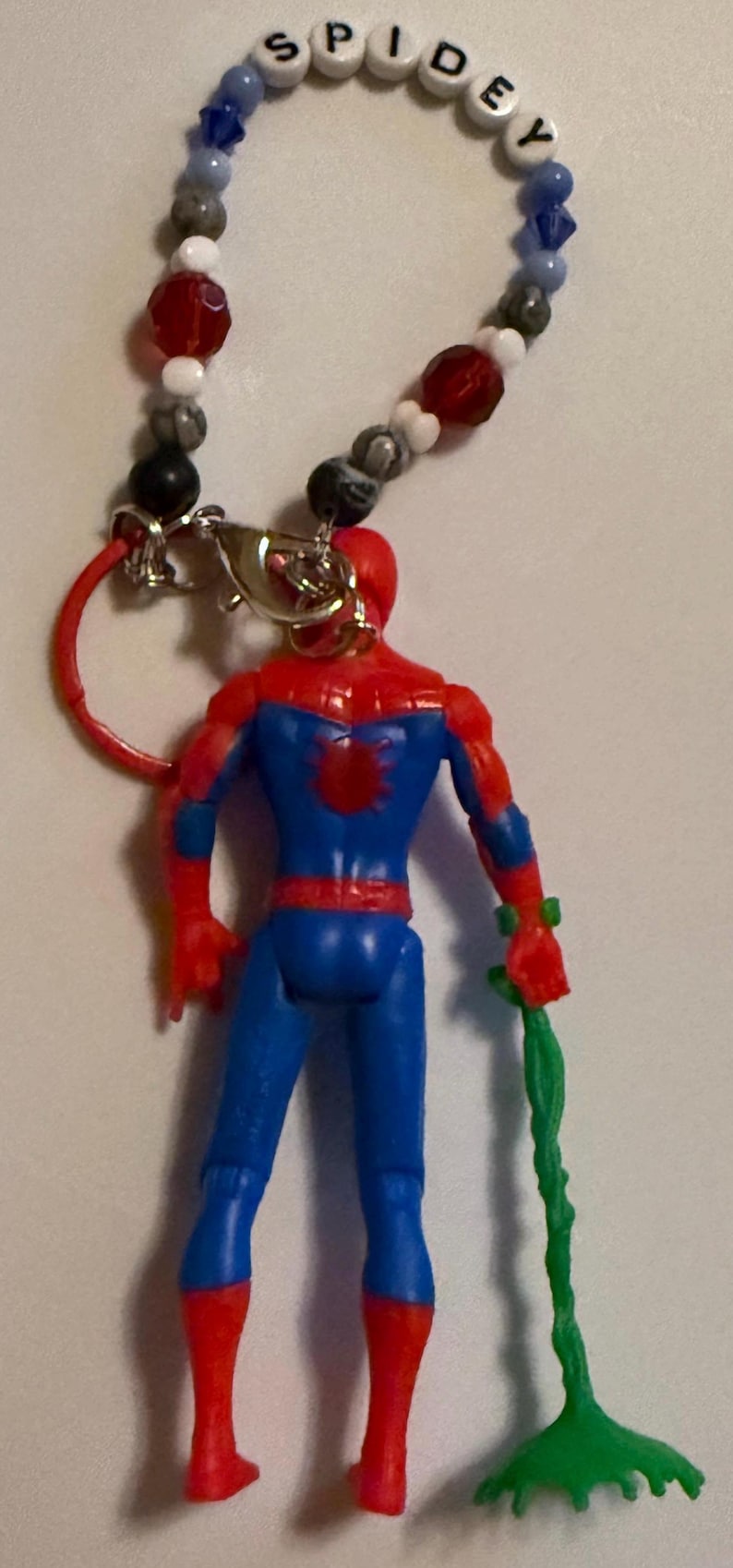 May include: A Spiderman figurine with a beaded lanyard that spells "SPIDEY". The figurine is red and blue, holding a green web-like accessory. The lanyard has blue, white, red, and black beads.