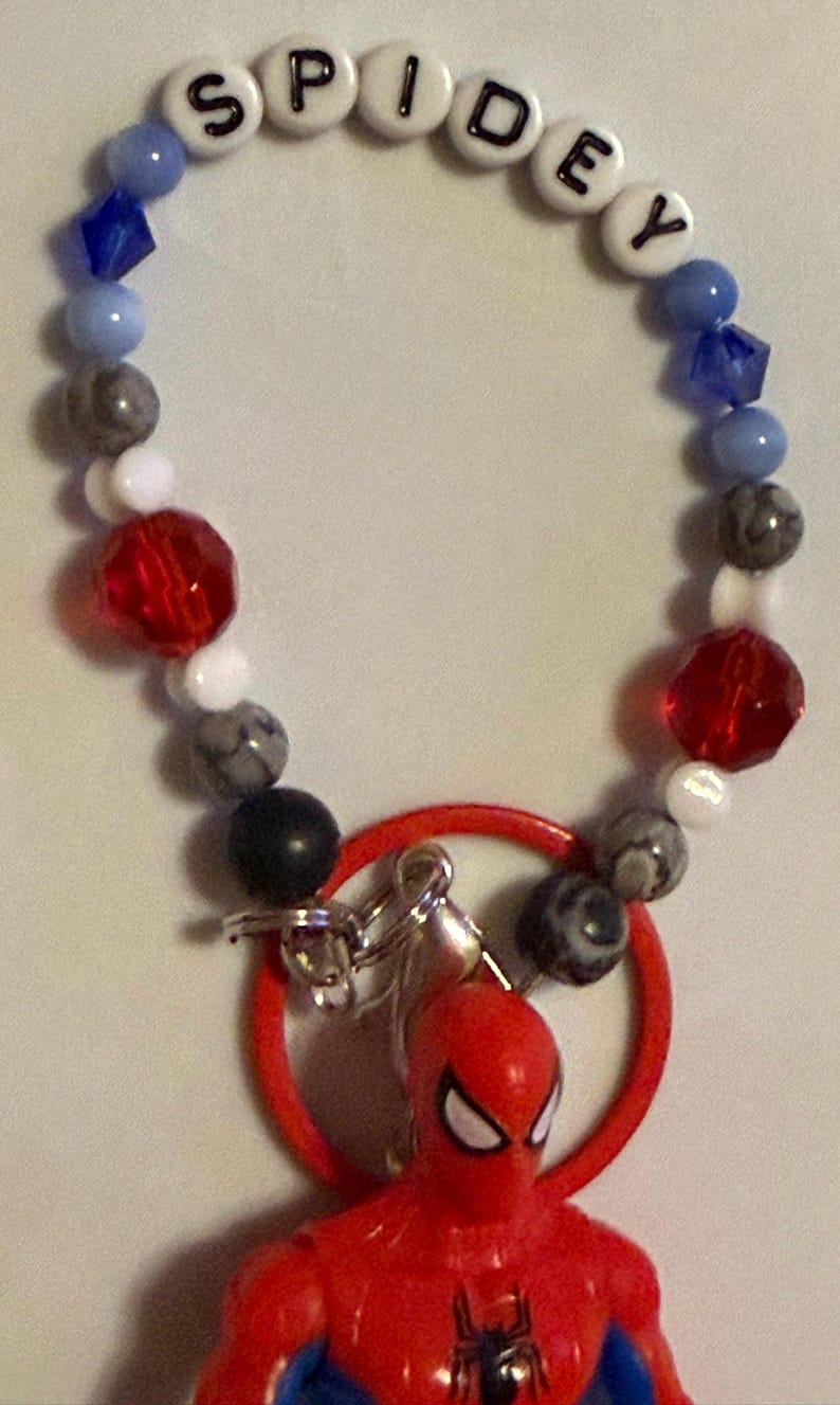 May include: A Spidey-themed keychain featuring a red Spider-Man figure, a red ring, and a beaded bracelet. The bracelet has beads in various colors, including blue, white, gray, and red, with the name "SPIDEY" spelled out.