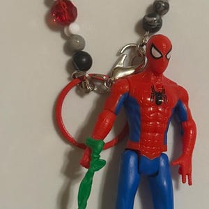 May include: A Spider-Man action figure with a red and blue suit, holding a green web. The figure is attached to a beaded keychain spelling "SPIDEY". The beads are various colors including red, white, gray, and blue.