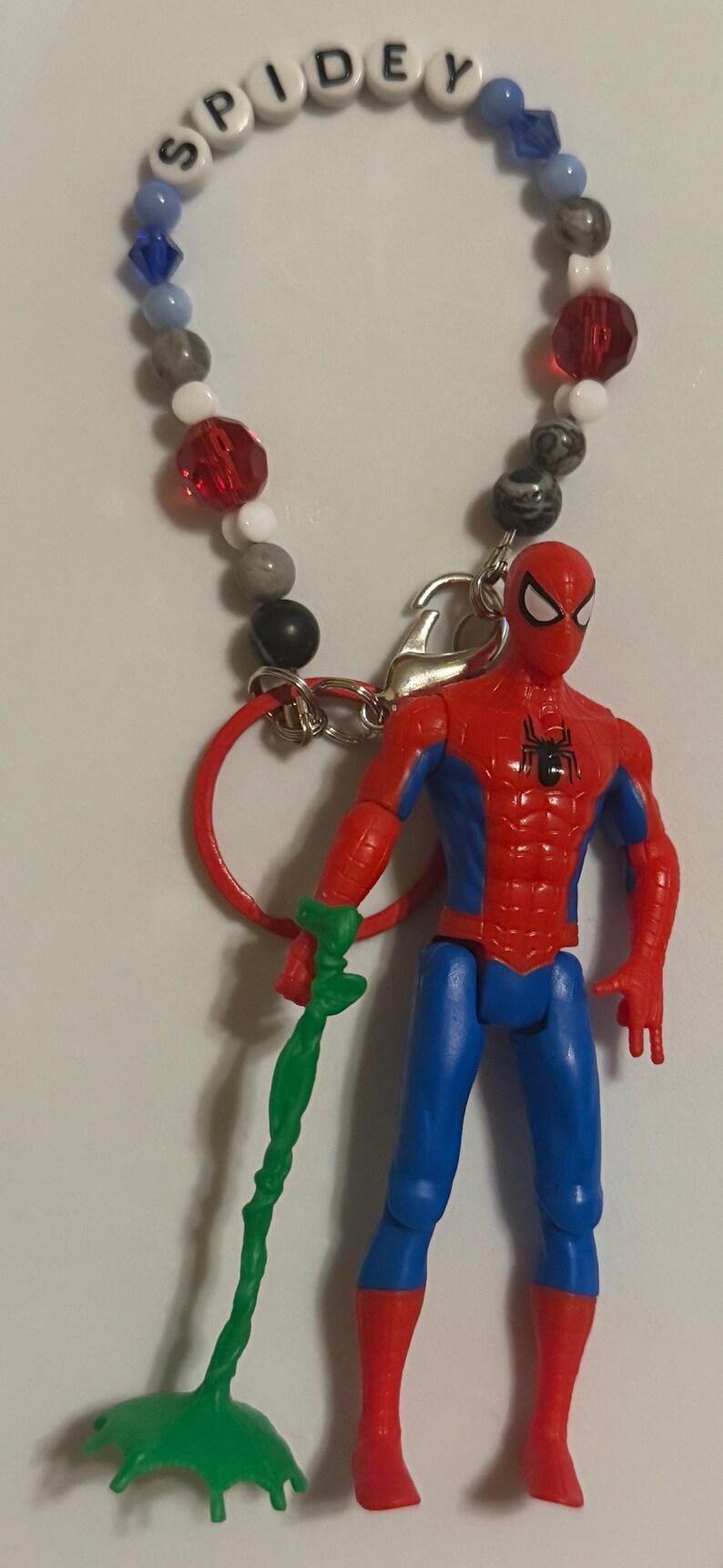 May include: A Spider-Man action figure with a red and blue suit, holding a green web. The figure is attached to a beaded keychain spelling "SPIDEY". The beads are various colors including red, white, gray, and blue.
