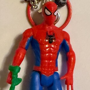 May include: A red, blue, and black Spider-Man action figure with a green accessory. The figure is attached to a red ring with a silver clasp and a beaded necklace. The figure has a black spider emblem on the chest.
