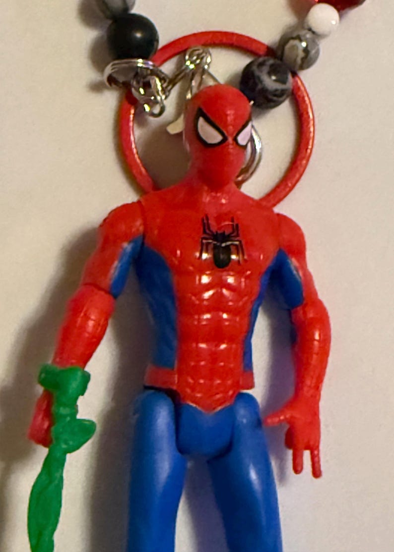 May include: A red, blue, and black Spider-Man action figure with a green accessory. The figure is attached to a red ring with a silver clasp and a beaded necklace. The figure has a black spider emblem on the chest.