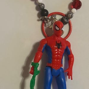 May include: A Spiderman action figure with a green accessory, attached to a beaded bracelet spelling "SPIDEY". The figure is red and blue, with a red ring and a silver clasp.