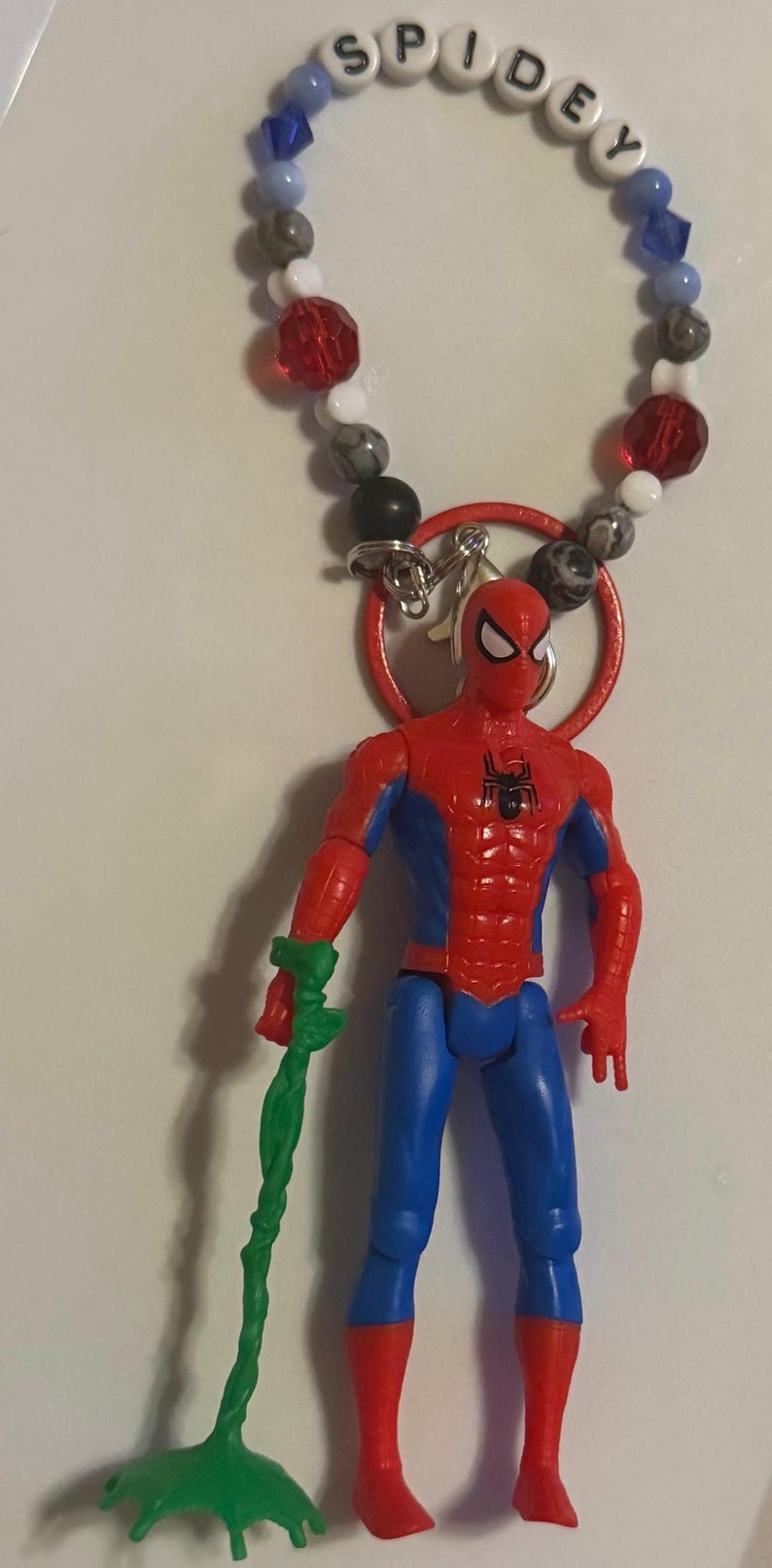 May include: A Spiderman action figure with a green accessory, attached to a beaded bracelet spelling "SPIDEY". The figure is red and blue, with a red ring and a silver clasp.