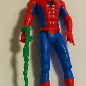 May include: A Spider-Man action figure with a red and blue costume, holding a green, vine-like accessory. The figure has a black spider emblem on the chest and is attached to a beaded keychain.