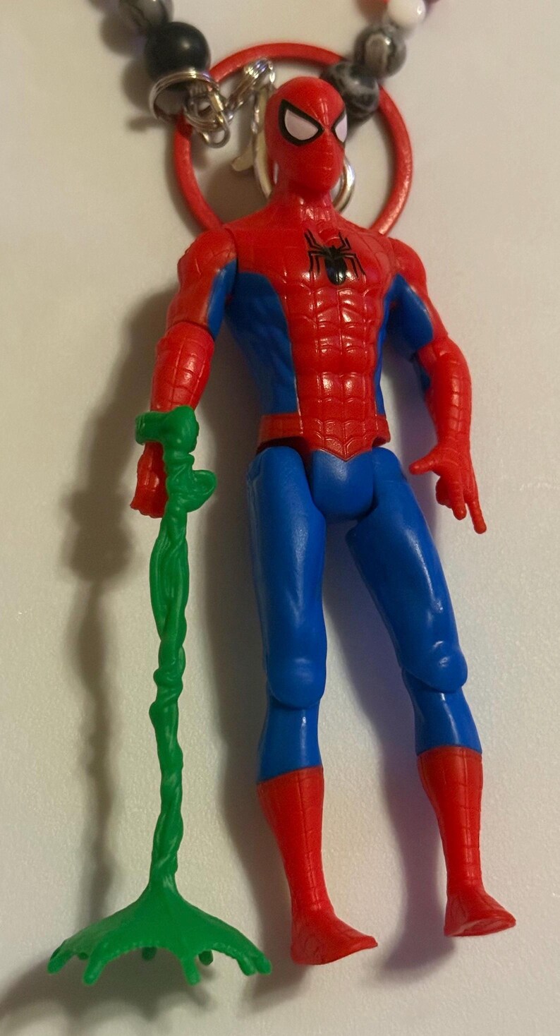 May include: A Spider-Man action figure with a red and blue costume, holding a green, vine-like accessory. The figure has a black spider emblem on the chest and is attached to a beaded keychain.