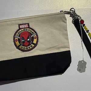May include: Beige canvas zip pouch featuring the Deadpool Marvel Collector Corps logo, with a black base. A black wrist strap with beads and the word "TACOS" is attached to the pouch.