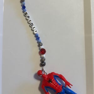 May include: A Spiderman keychain featuring a red and blue Spiderman figure with a green web. The keychain has a beaded strand with the word "SPIDEY" spelled out in white letters. The beads are red, white, gray, and blue.