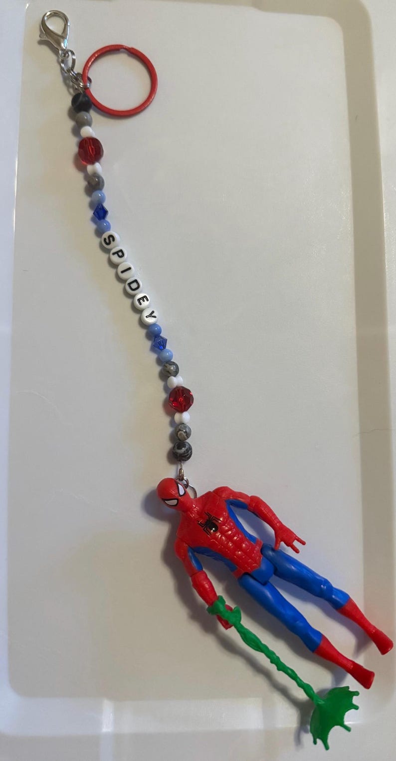 May include: A Spiderman keychain featuring a red and blue Spiderman figure with a green web. The keychain has a beaded strand with the word "SPIDEY" spelled out in white letters. The beads are red, white, gray, and blue.