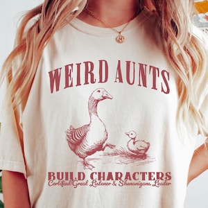 May include: A beige t-shirt with the text "Weird Aunts Build Characters Certified Great Listener & Shenanigans Leader" in red lettering. The shirt features a drawing of two geese.