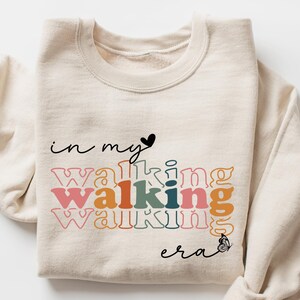 May include: A sand-colored sweatshirt with the text "in my walking era" in a layered, colorful design. The sweatshirt has long sleeves and a crew neck. A small black heart and butterfly accent the text.