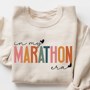 May include: A beige sweatshirt with the text "in my MARATHON era" printed in rainbow colors. The text is in a cursive font and has a small heart symbol next to the word "my".