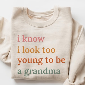Peut inclure: Sweat-shirt couleur sable avec le texte "i know i look too young to be a grandma" en rose, orange et vert. Le sweat-shirt est plié sur une surface blanche.