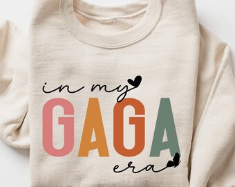 Può includere: Felpa color sabbia con la scritta "in my GAGA era" in nero, rosa, arancione e verde. La felpa ha un girocollo e maniche lunghe. La parola "sand" è in un rettangolo giallo in basso a sinistra.