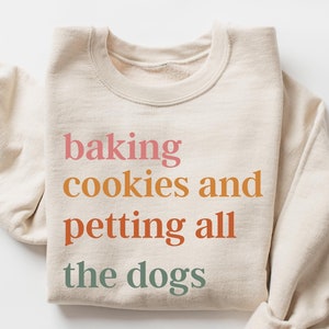 May include: A beige sweatshirt with the text "baking cookies and petting all the dogs" printed in a gradient of pink, orange, yellow, and green.
