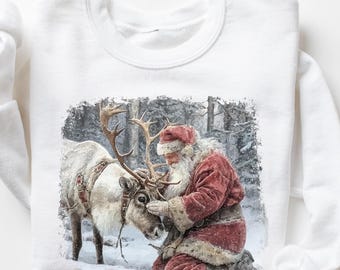 May include: White long-sleeved sweatshirt featuring a winter scene with Santa Claus and a reindeer. Santa is wearing a red suit and hat, kneeling next to the reindeer. The background shows a snowy forest.